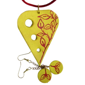 Souprava z kůže s červenými lístky. Přívěsek tvaru písmene V s otvory po jedné straně a kruhové náušnice vystřižené z kůže a malované žlutou akrylovou barvou s červenou kresbou lístečků jsou zavěšené na háčcích a bižuterním očku zlaté barvy.