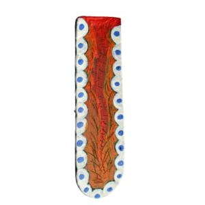 Malovaná brož. Brož ve tvaru dlouhého úzkého písmene U je vystřižená z kůže a malovaná akrylovou barvou. Po obvodu jsou bílé obloučky s modré puntíky a ve středu hrubá, plastická oranžová kresba. Ze spodní strany je pomocí průhledného plastu připevněný spínací špendlík zlaté barvy.