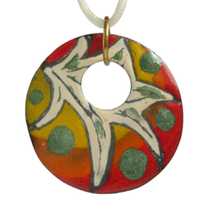 Smaltovaný mexický přívěsek. Přívěsek kruhového tvaru s větším kulatým otvorem s ostrou béžovou kresbou a zelenými puntíky na žlutooranžovočerveném podkladu na ručně opracovaném nerezovém plíšku. Je zavěšený za velké bižuterní očko zlaté barvy.
