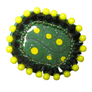 Opletená smaltovaná brož se žlutými puntíky. Základový ručně opracovaný nerezový plíšek oválného tvaru se zelenou smaltovou vrstvou a žlutými puntíky je opletený zelenými černými a žlutými korálky je podšitá zelenou plstí. Zespodu je přišitý zavírací špendlík zlaté barvy.