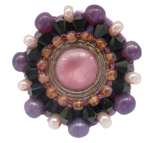 Fialová korálková brož. Brož vyrobená z růžového jabloneckého knoflíku se stříbrným okrajem, který je opletený pomocí měděného drátku a korálků v černé, hnědé, fialové a růžové barvě. Je podšitá fialovou plstí. Ze spodní strany je přišitý špendlík v černé barvě.