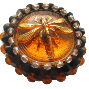 Korálková brož s vážkou. Brož vyrobená z oranžového jabloneckého knoflíku se zlatým reliéfem vážky, který je opletený pomocí měděného drátku a korálků v černé, oranžové a růžové barvě. Je podšitá černou plstí. Ze spodní strany je pomocí průhledného plastu připevněný špendlík v černé barvě.