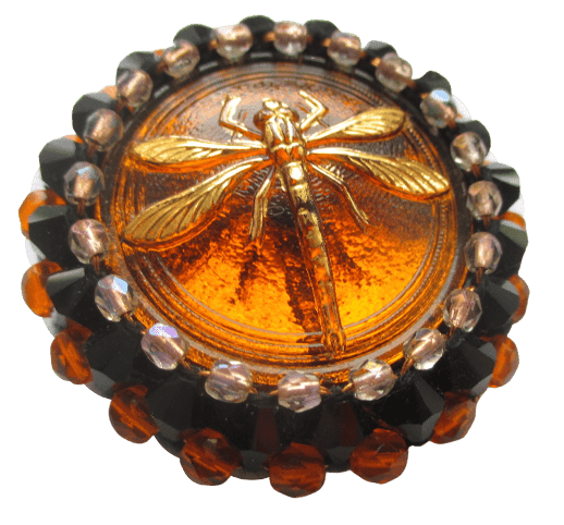 Korálková brož s vážkou. Brož vyrobená z oranžového jabloneckého knoflíku se zlatým reliéfem vážky, který je opletený pomocí měděného drátku a korálků v černé, oranžové a růžové barvě. Je podšitá černou plstí. Ze spodní strany je pomocí průhledného plastu připevněný špendlík v černé barvě.