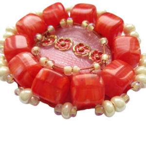 Korálková romantická brož. Brož vyrobená z oválného růžového jabloneckého knoflíku s reliéfem zlatočervených květin, který je opletený pomocí měděného drátku a korálků v červené a perleťové barvě. Je podšitá červenou plstí. Ze spodní strany je přišitý špendlík ve stříbrné barvě.