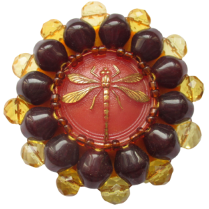 Korálková brož s vážkou. Brož vyrobená z červeného jabloneckého knoflíku se zlatým reliéfem vážky, který je opletený pomocí měděného drátku a korálků v hnědé a žluté barvě. Je podšitá žlutou plstí. Ze spodní strany je přišitý špendlík ve zlaté barvě.